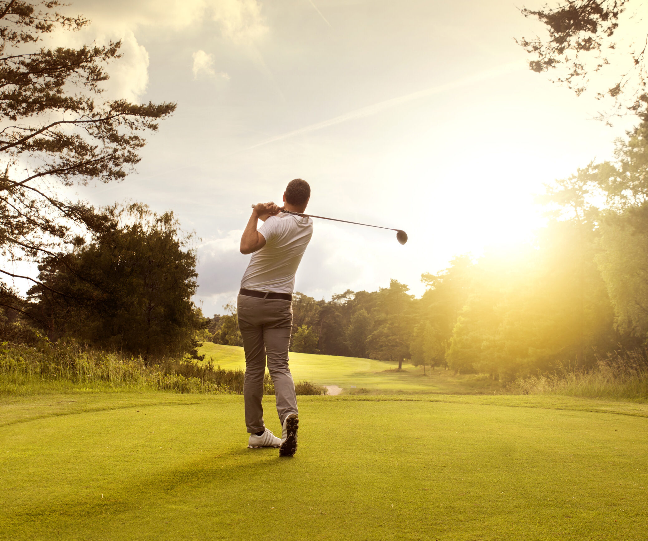 Man playing golf at golf course in Park City hosted by Luxe Haus. There is a sunset view and the man is swinging a club wearing a white shirt and beige pants