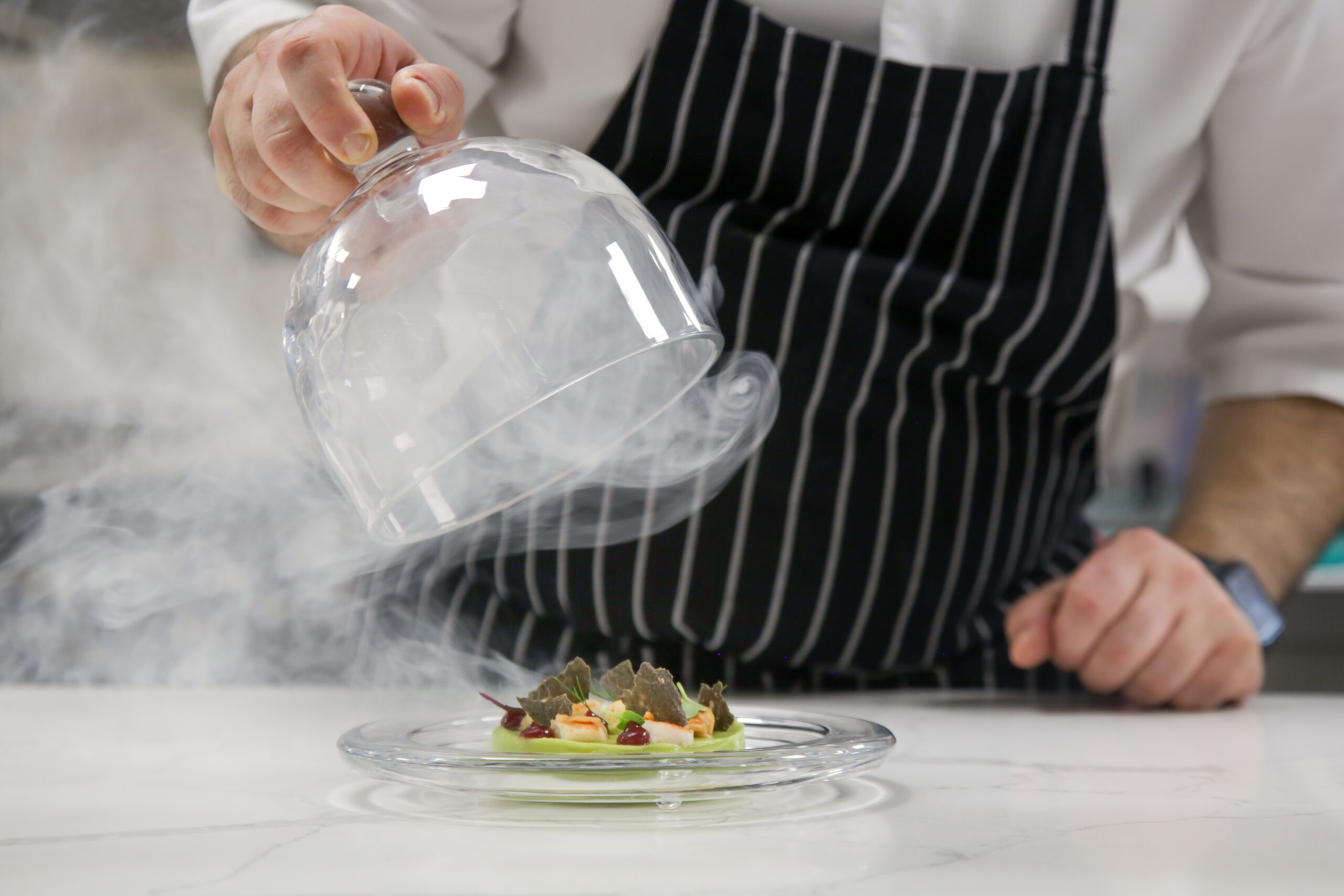 Chef's hand lifts up glass cloche from a plate with hot food and moving smoke at the restaurant. Exquisite dish, creative restaurant meal concept, haute couture food in Park City, hosted by Luxe Haus