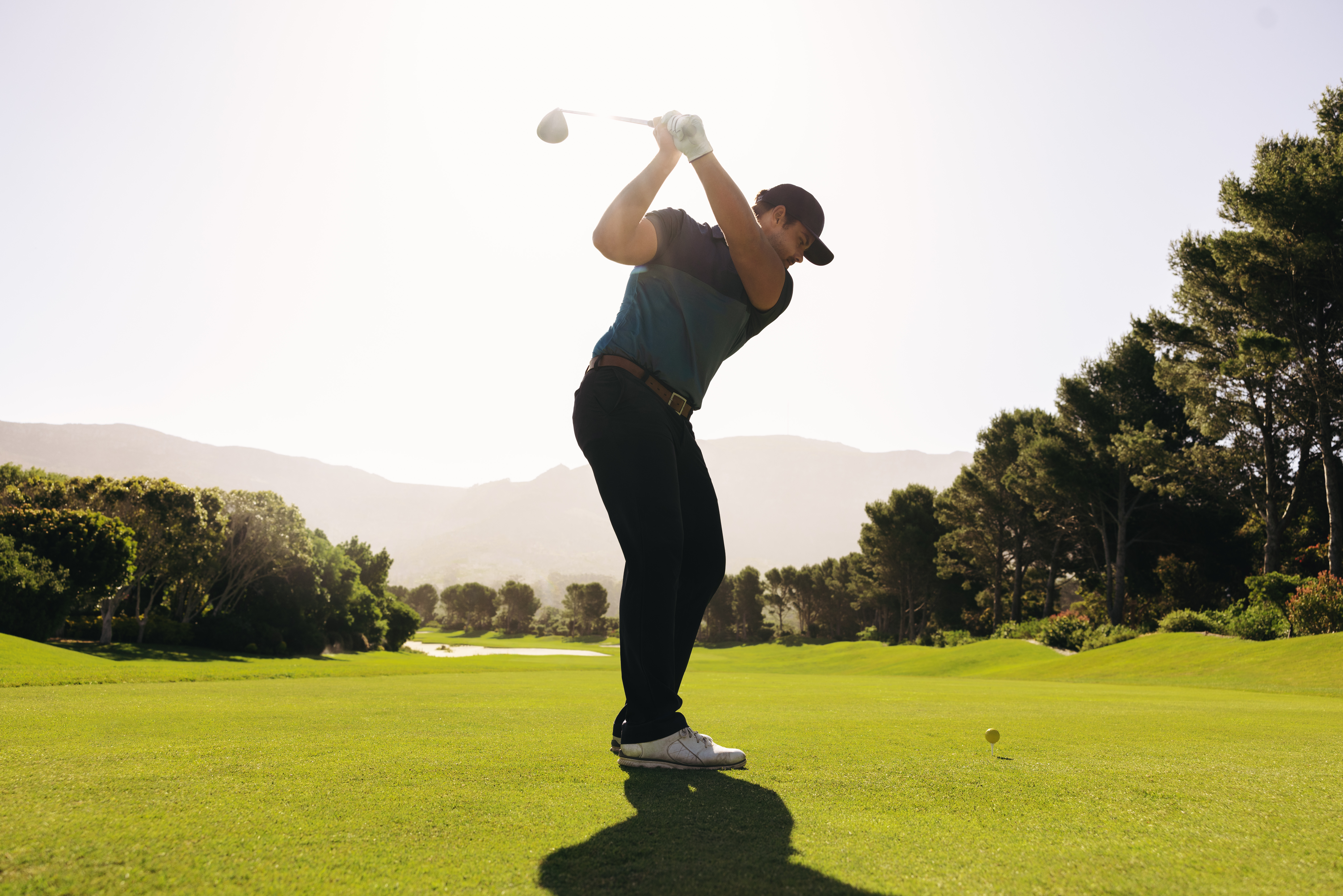A man golfer skillfully swings his club during a fairway shot in park city, with a golf ball in sight, surrounded by vibrant greenery under a clear sunny sky.