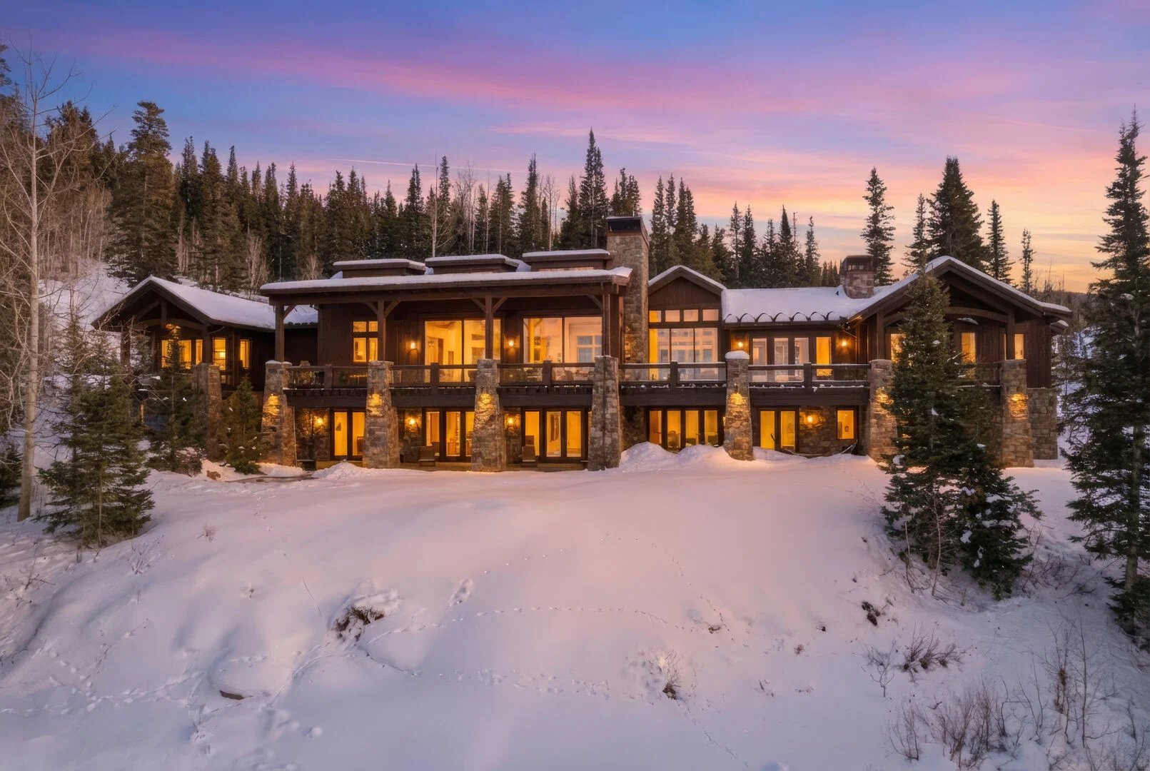 A view of snow dance in park city at sundown, the sky is glowing pink and the mountains glisten with snow behind the home which is glowing from within with warm light