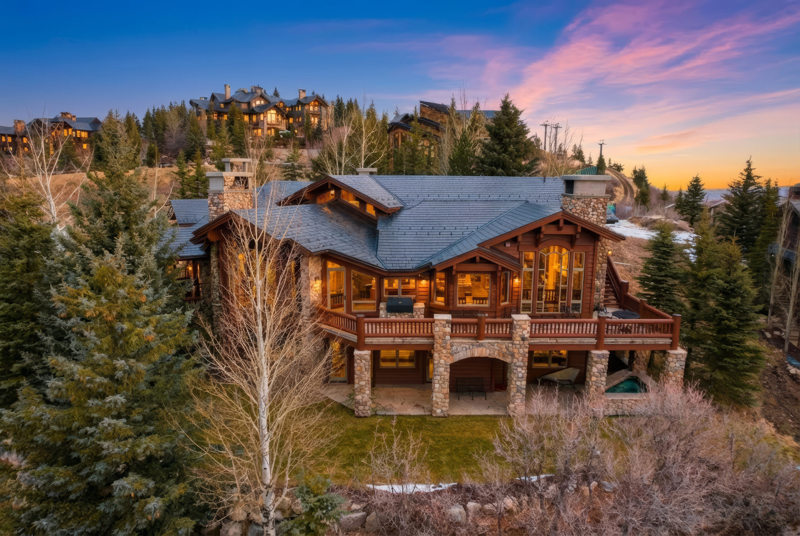 Summit View Lodge at Deer Crest at sunset. the lights are glowing from inside a stately log cabin lodge with the view of the mountains of park city in the background